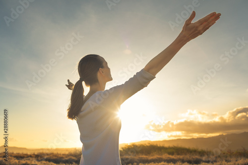 Young female looking up to the sunset sky with arms stretched out. Happiness and freedom outdoors concept. 