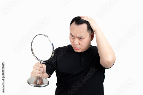 Portrait of serious young Asian bald man with alopecia, checking hairline while looking at the mirror isolated on white background. Hair loss, hair transplant concept.