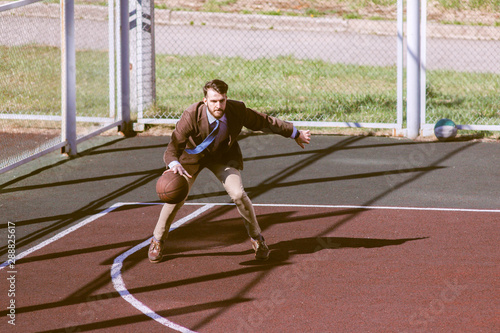 Stylish bearded handsome hipster guy in a trendy suit in a jacket and tie on a basketball court playing basketball. Fashionable handsome man with a ball in his hand, top view. Business concept, sport.