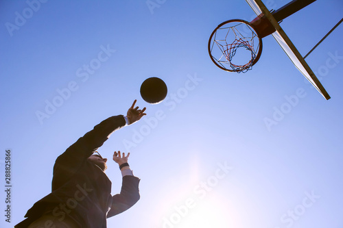 Bottom view of a guy in a business suit throwing a basketball into the basket on the sports field. The combination of office work and sports. Businessman playing basketball against the sky.