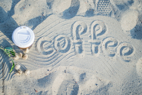 The inscription "coffee" on the sand. Paper cup with coffee. Bright summer good morning. Brown paper cup with plastic white lid.