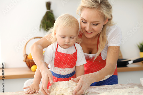 Little girl and her blonde mom in red aprons playing and laughing while kneading the dough in kitchen. Homemade pastry for bread, pizza or bake cookies. Family fun and cooking concept