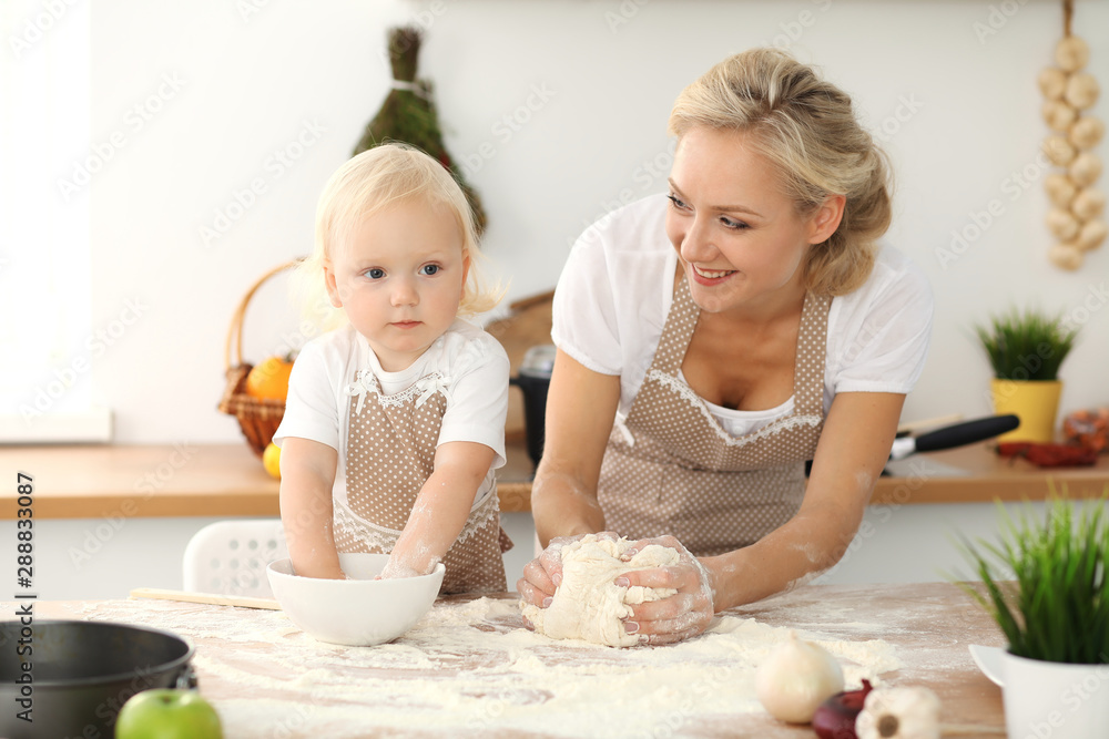 Little girl and her blonde mom in beige aprons playing and laughing while kneading the dough in kitchen. Homemade pastry for bread, pizza or bake cookies. Family fun and cooking concept Little girl and her blonde mom in beige aprons playing and laughing while kneading the dough in kitchen. Homemade pastry for bread, pizza or bake cookies. Family fun and cooking concept