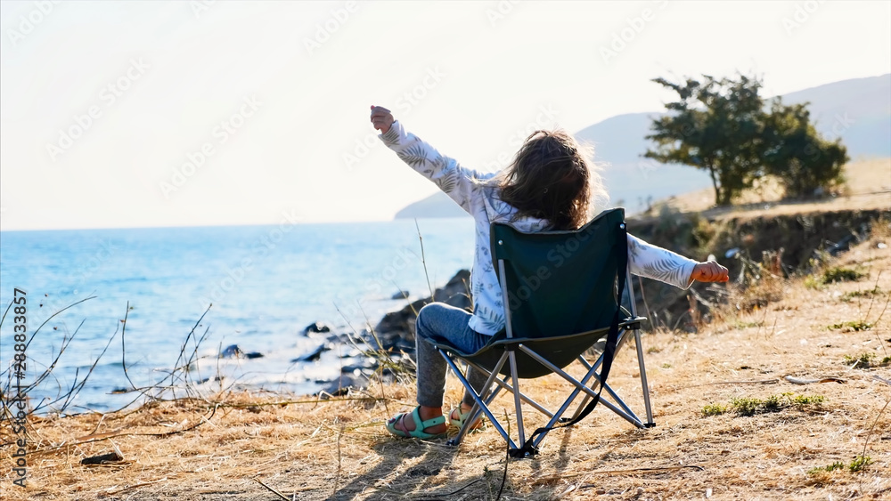 Rear view of little child girl sitting on folding chair on sea shore at early morning in windy weather and stretching in slow motion 