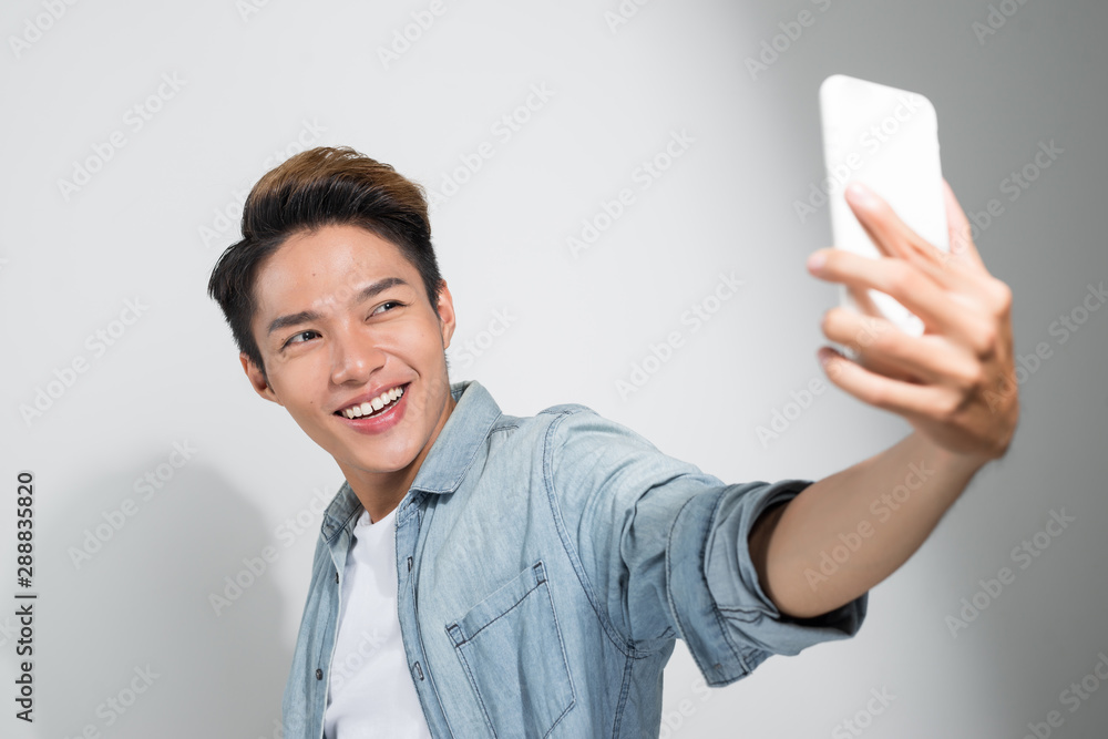 Studio shot of a smiling Asian young man making selfie with the smartphone while standing over white background Studio shot of a smiling Asian young man making selfie with the smartphone while standing over white background