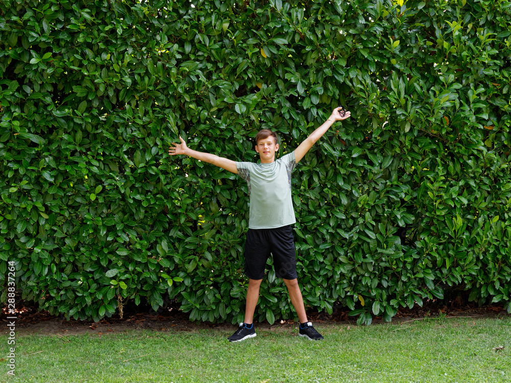 teenager stands on a background of green hedges.