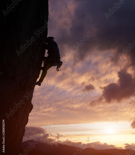 Male climber silhouette overcoming difficult path to top on challenging cliff in nightfall. Rock climbing. Cloudy sky with cumulonimbus over mountain peaks on background. Low angle view. Copy space