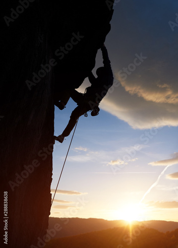 Male silhouette rock climbing, doing next step at sunrise in mountains. Rock climber hanging on cliff above glowing horizon under big clouds. Side view. Dangerous, extreme, endurance, victory concept