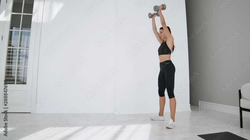 Strong and Fit Beautiful Girl in an Athletic Top is Doing Squat Exercises in Her Bright and Spacious Living Room with Minimalistic Interior. Strong and Fit Beautiful Girl in an Athletic Top is Doing Squat Exercises in Her Bright and Spacious Living Room with Minimalistic Interior.