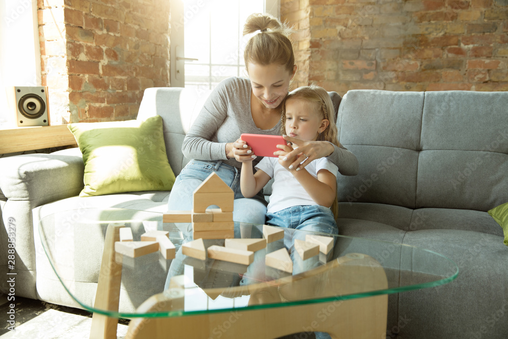 Female caucasian teacher and little girl, or mom and daughter. Homeschooling. Sitting on the sofa with wooden constructor, watching guide, talking and having fun. Education, school, studying concept. Female caucasian teacher and little girl, or mom and daughter. Homeschooling. Sitting on the sofa with wooden constructor, watching guide, talking and having fun. Education, school, studying concept.