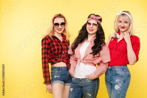 Studio shot of trendy girls with head bows, sunglasses wearing trendy hipster clothes and smiling at camera. Isolate on yellow background.