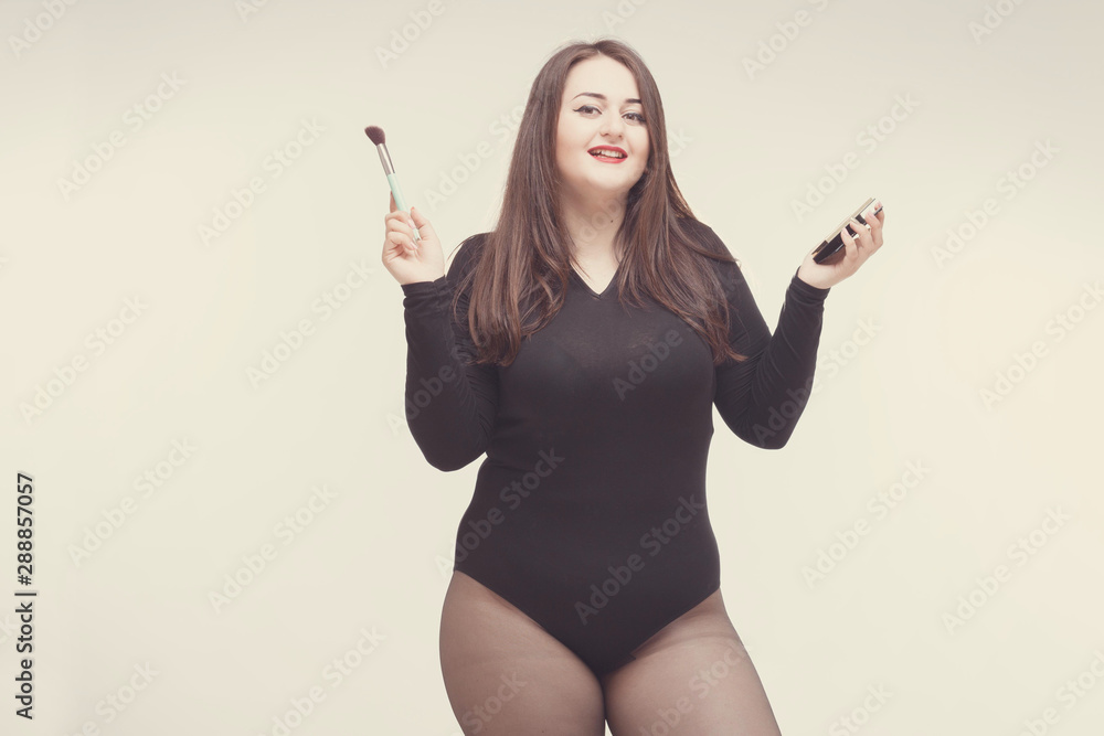 Model plus size with sweet donut, happy smiling girl posing with cosmetic, doing makeup for her self with brush in black bodysuit. XXL female on light background with space for text Model plus size with sweet donut, happy smiling girl posing with cosmetic, doing makeup for her self with brush in black bodysuit. XXL female on light background with space for text