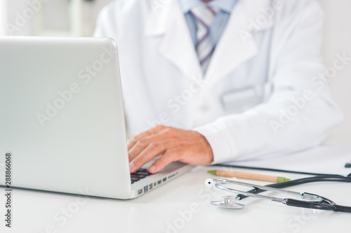 Senior doctor at his office in hospital working close-up using laptop typing front view
