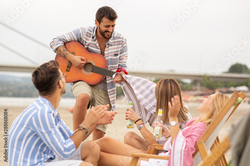 Group of friends sitting at the beach and play guitar and singing.