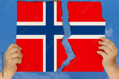 woman holds in both hands against a blue sky torn in two cardboard with seared edges depicting the national flag of Norway, concept of state crisis, renunciation of citizenship, destruction