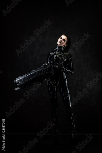 Portrait of a young woman in latex jumpsuit holding a big futuristic weapon in her hands