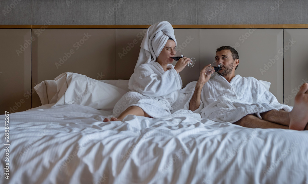 Fototapeta premium Happy lovely couple wearing white bathrobes smiling to each other and drinking wine in bed at hotel room. Enjoying honeymoon in luxury resort