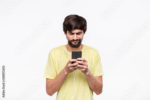 Handsome brunet bearded man with whiskers in a yellow tee holding smartphone with connected wired headphones expressing emotion of distrust isolated over white background. Receiving unexpected news. photo