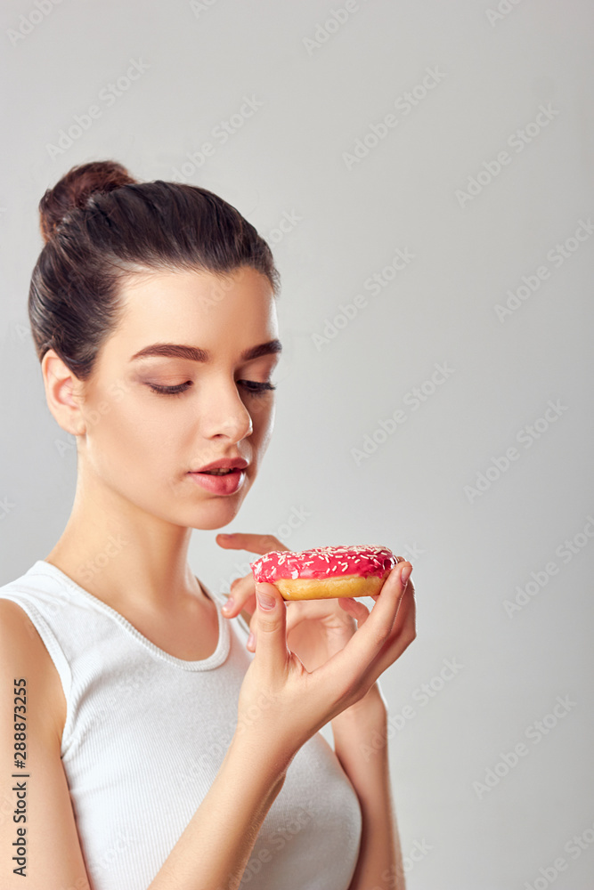 Close up portrait of a hungry girl eating donuts isolated over white background.Brunette girl holds and tastes a pink donut. Sweets are unhealthy junk food. Dieting. Healthy Eating. Close up portrait of a hungry girl eating donuts isolated over white background.Brunette girl holds and tastes a pink donut. Sweets are unhealthy junk food. Dieting. Healthy Eating.