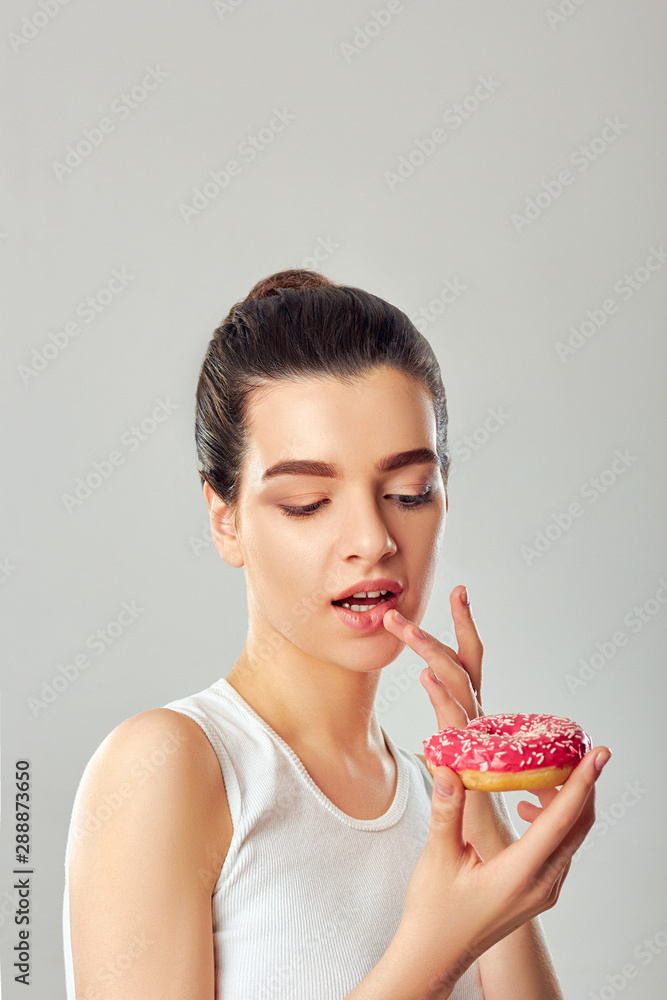 Beauty model girl holds a pink donut. Diet. Funny. Brunette woman tastes and eating a donut Beauty model girl holds a pink donut. Diet. Funny. Brunette woman tastes and eating a donut