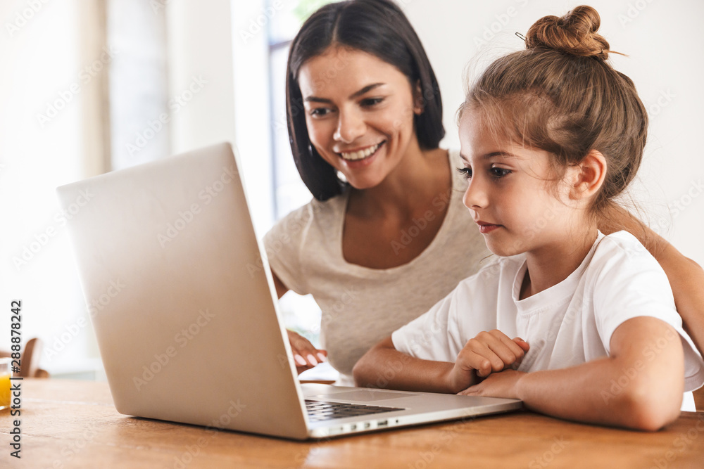 Image of attractive family woman and her little daughter smiling and using laptop computer together in apartment Image of attractive family woman and her little daughter smiling and using laptop computer together in apartment