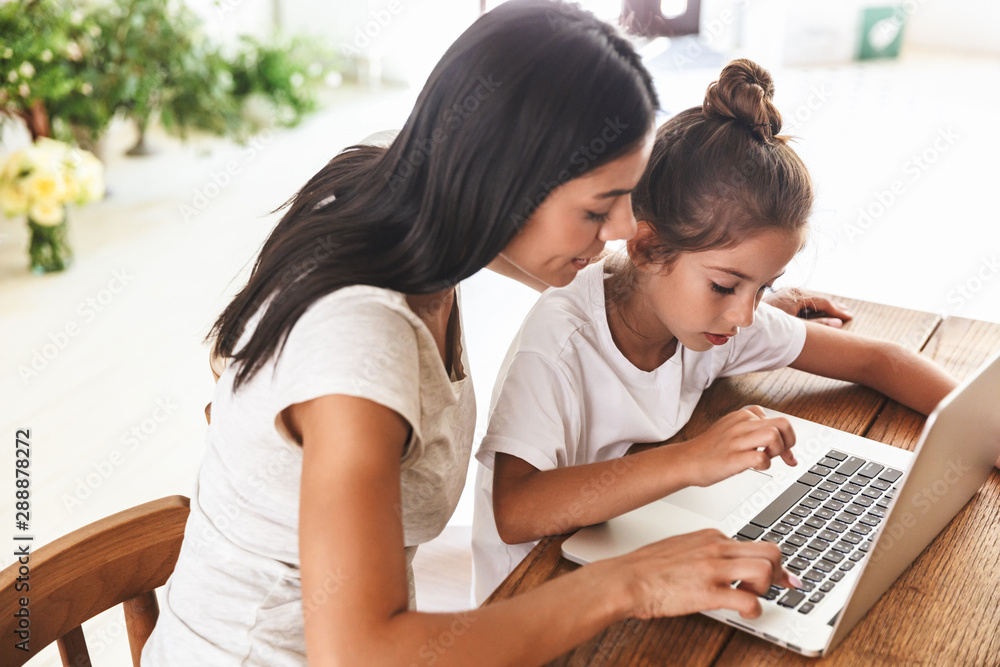 Image of pretty family woman and her little daughter smiling and using laptop computer together in apartment