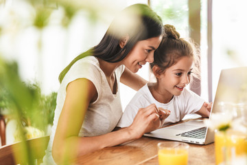 Image of gorgeous family woman and her little daughter smiling and using laptop computer together in apartment