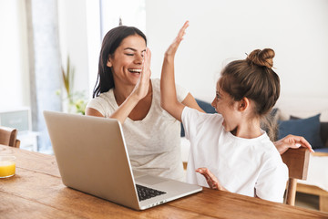 Image of excited family woman and her little daughter giving high five and using laptop computer together in apartment