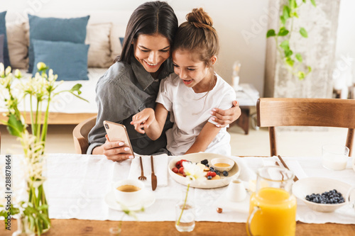 Image of caucasian family mother and little daughter using cellphone while having breakfast at home in morning