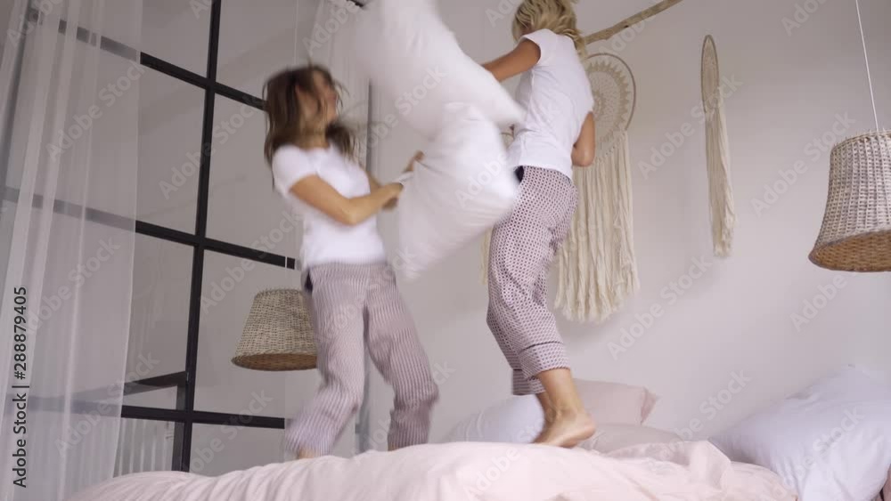 Two adorable fun girlfriends in the same pajamas jumping on the bed laughing and fighting with pillows in the light modern room. Hen-party. Leisure at home Two adorable fun girlfriends in the same pajamas jumping on the bed laughing and fighting with pillows in the light modern room. Hen-party. Leisure at home