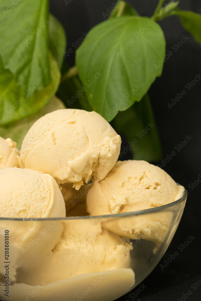 Homemade mango ice cream in yellow in a glass clear deep plate on a black background and green plant Homemade mango ice cream in yellow in a glass clear deep plate on a black background and green plant