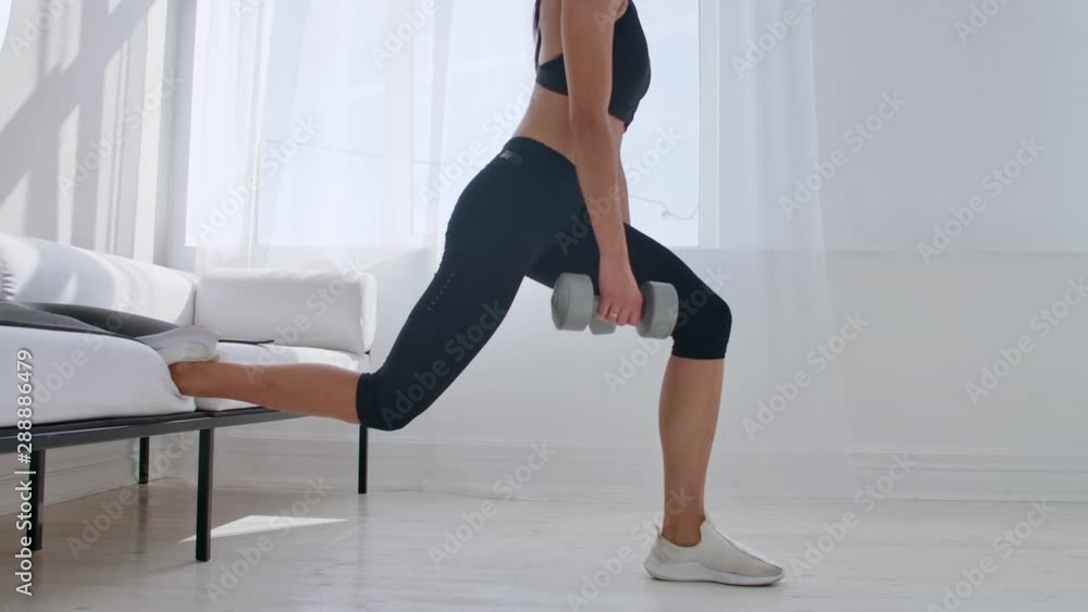 Brunette in black sportswear in a white apartment makes a split squat with dumbbells in her hands leaning on the sofa with her foot Brunette in black sportswear in a white apartment makes a split squat with dumbbells in her hands leaning on the sofa with her foot