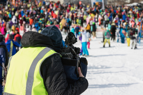 Unrecognizable sports operator with a video camera, back to us, shooting people outdoors, sports event on the ski slope. Behind the scenes