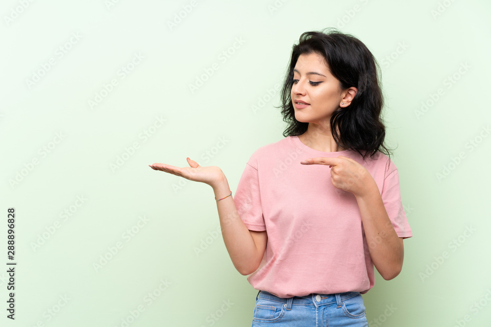 Young woman over isolated green background holding copyspace imaginary on the palm to insert an ad Young woman over isolated green background holding copyspace imaginary on the palm to insert an ad
