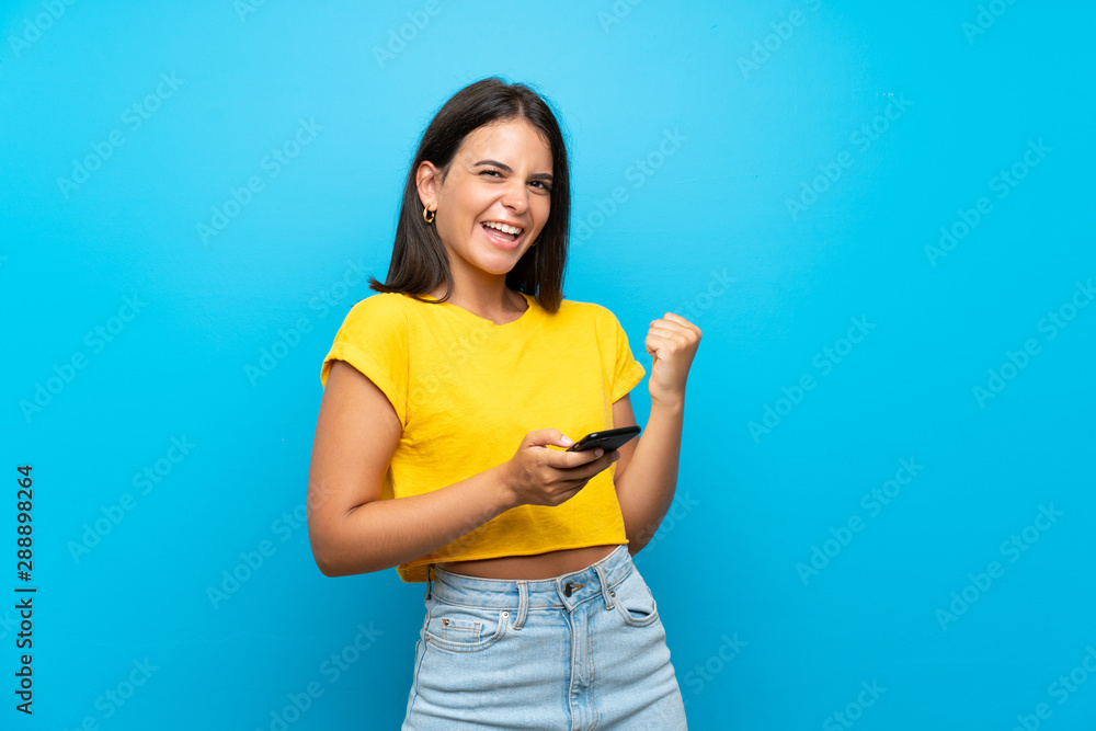 Young girl over isolated blue background with phone in victory position Young girl over isolated blue background with phone in victory position