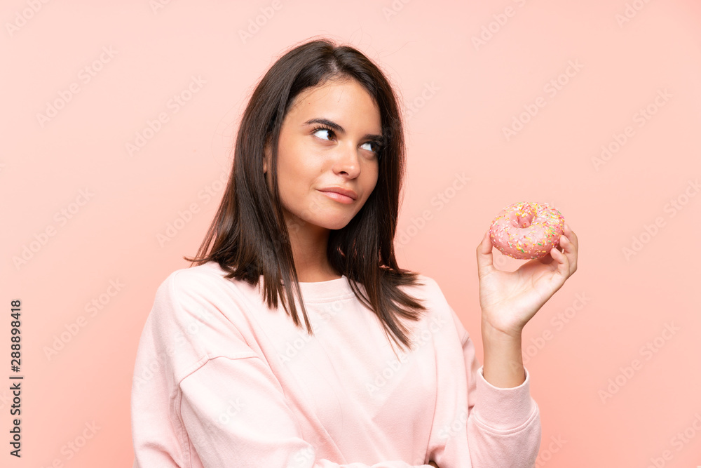 Young girl holding a donut over isolated pink background making doubts gesture while lifting the shoulders Young girl holding a donut over isolated pink background making doubts gesture while lifting the shoulders