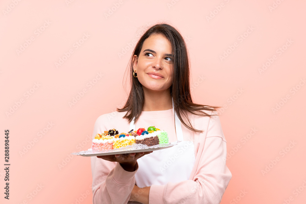 Young girl holding lots of different mini cakes over isolated background looking up while smiling Young girl holding lots of different mini cakes over isolated background looking up while smiling