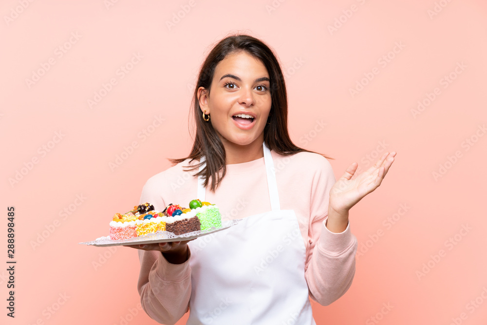 Young girl holding lots of different mini cakes over isolated background with shocked facial expression Young girl holding lots of different mini cakes over isolated background with shocked facial expression