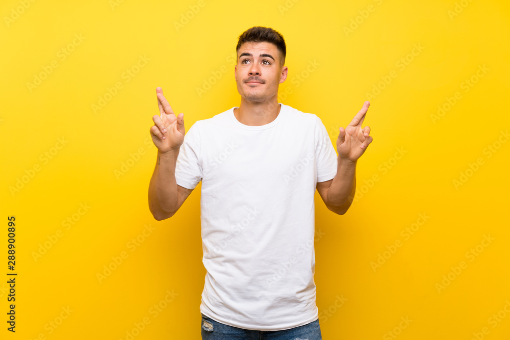 Young handsome man over isolated yellow background with fingers crossing and wishing the best Young handsome man over isolated yellow background with fingers crossing and wishing the best