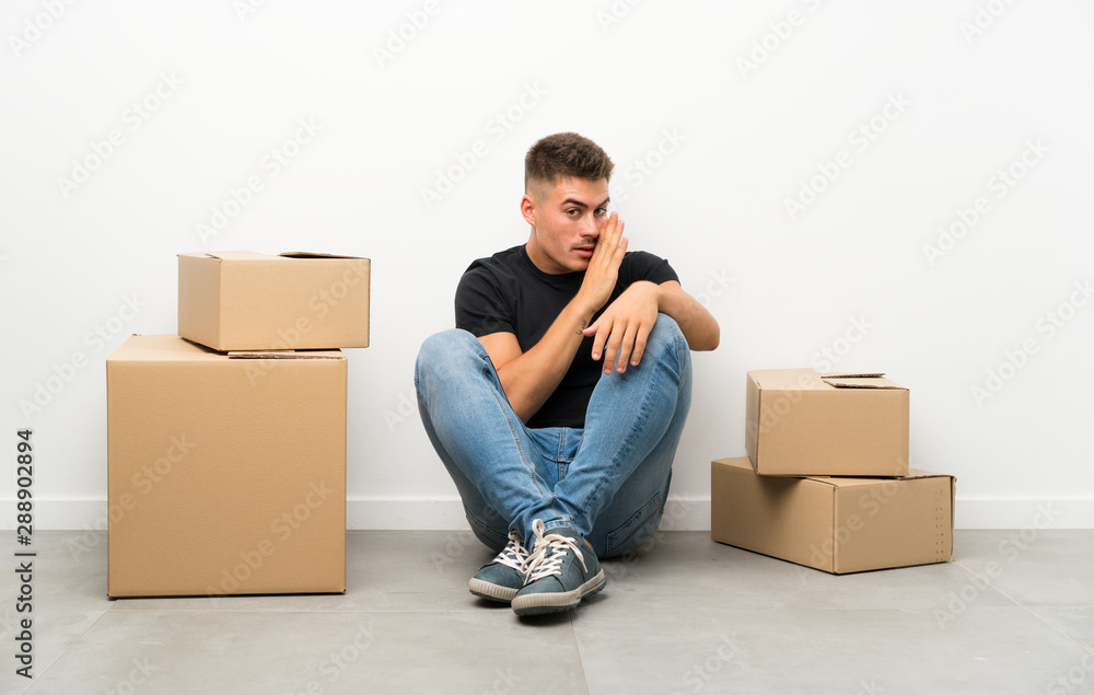 Handsome young man moving in new home among boxes whispering something Handsome young man moving in new home among boxes whispering something