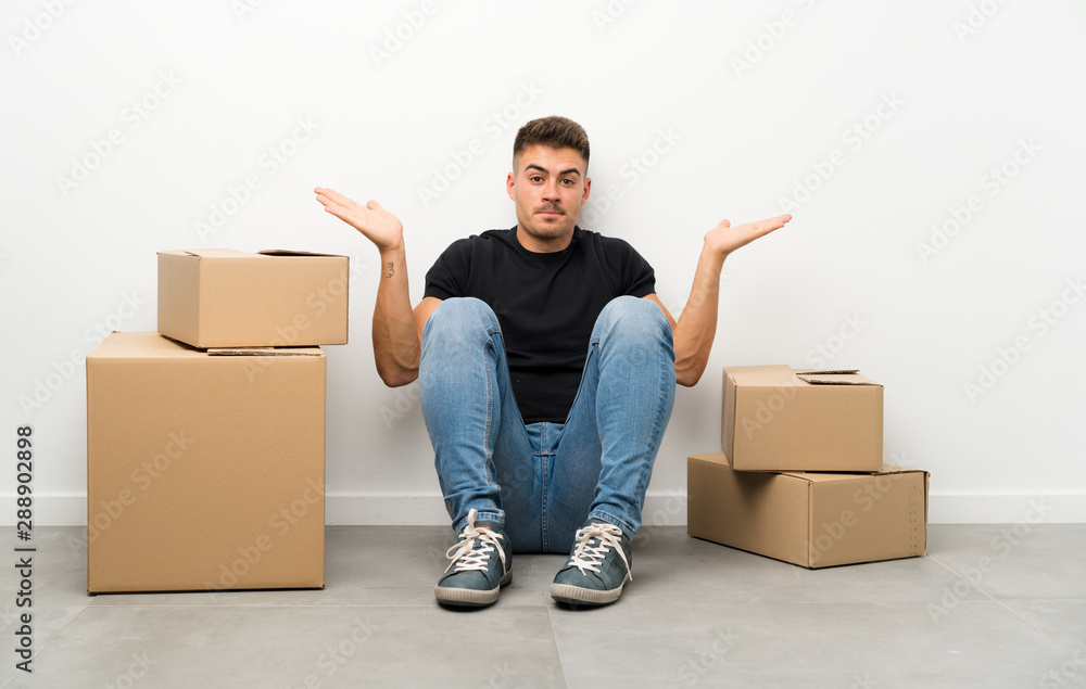 Handsome young man moving in new home among boxes having doubts with confuse face expression Handsome young man moving in new home among boxes having doubts with confuse face expression