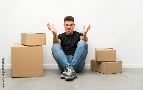 Handsome young man moving in new home among boxes unhappy and frustrated with something