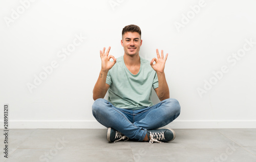 Young handsome man sitting on the floor showing an ok sign with fingers