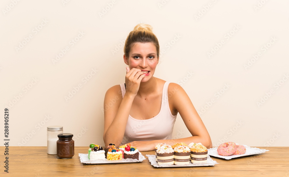 Young woman with lots of different mini cakes in a table nervous and scared Young woman with lots of different mini cakes in a table nervous and scared