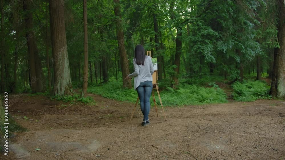 Girl draws a landscape, standing in front of an easel in a park. General plan beautiful women in jeans and a jacket on the background of the lake and tree trunks. Wide shot. Girl draws a landscape, standing in front of an easel in a park. General plan beautiful women in jeans and a jacket on the background of the lake and tree trunks. Wide shot.