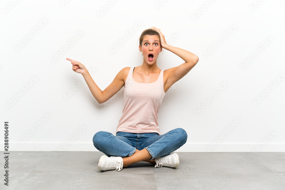 Young blonde woman sitting on the floor surprised and pointing finger to the side Young blonde woman sitting on the floor surprised and pointing finger to the side