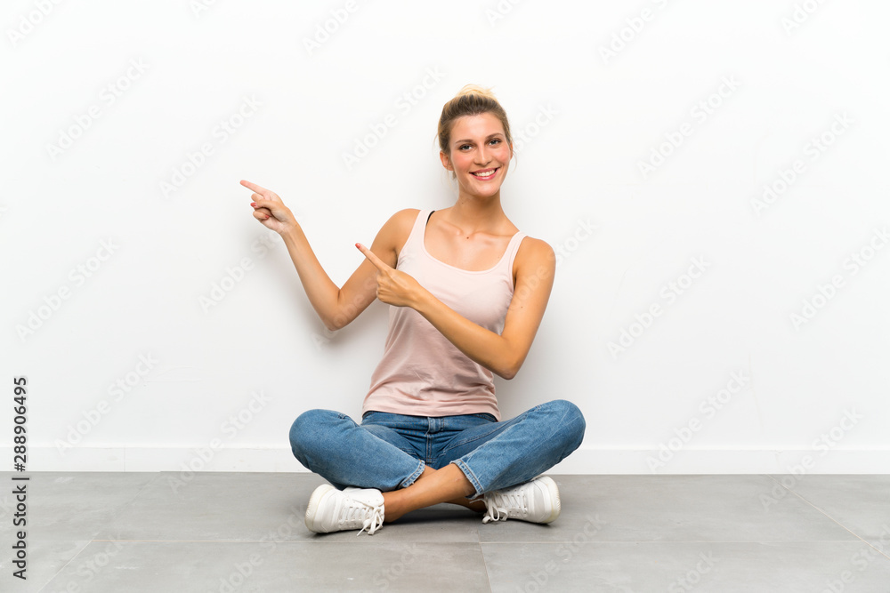Young blonde woman sitting on the floor pointing finger to the side Young blonde woman sitting on the floor pointing finger to the side