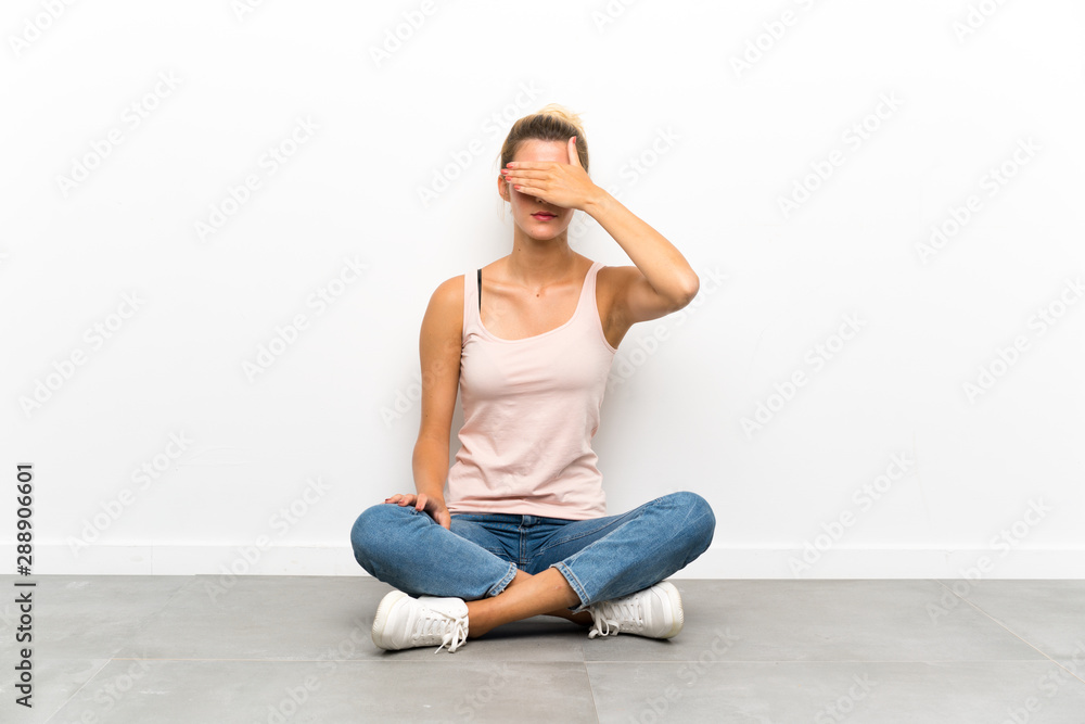 Young blonde woman sitting on the floor covering eyes by hands