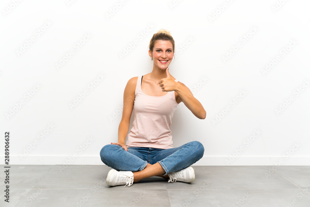 Young blonde woman sitting on the floor giving a thumbs up gesture