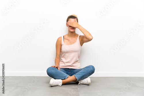 Young blonde woman sitting on the floor covering eyes by hands